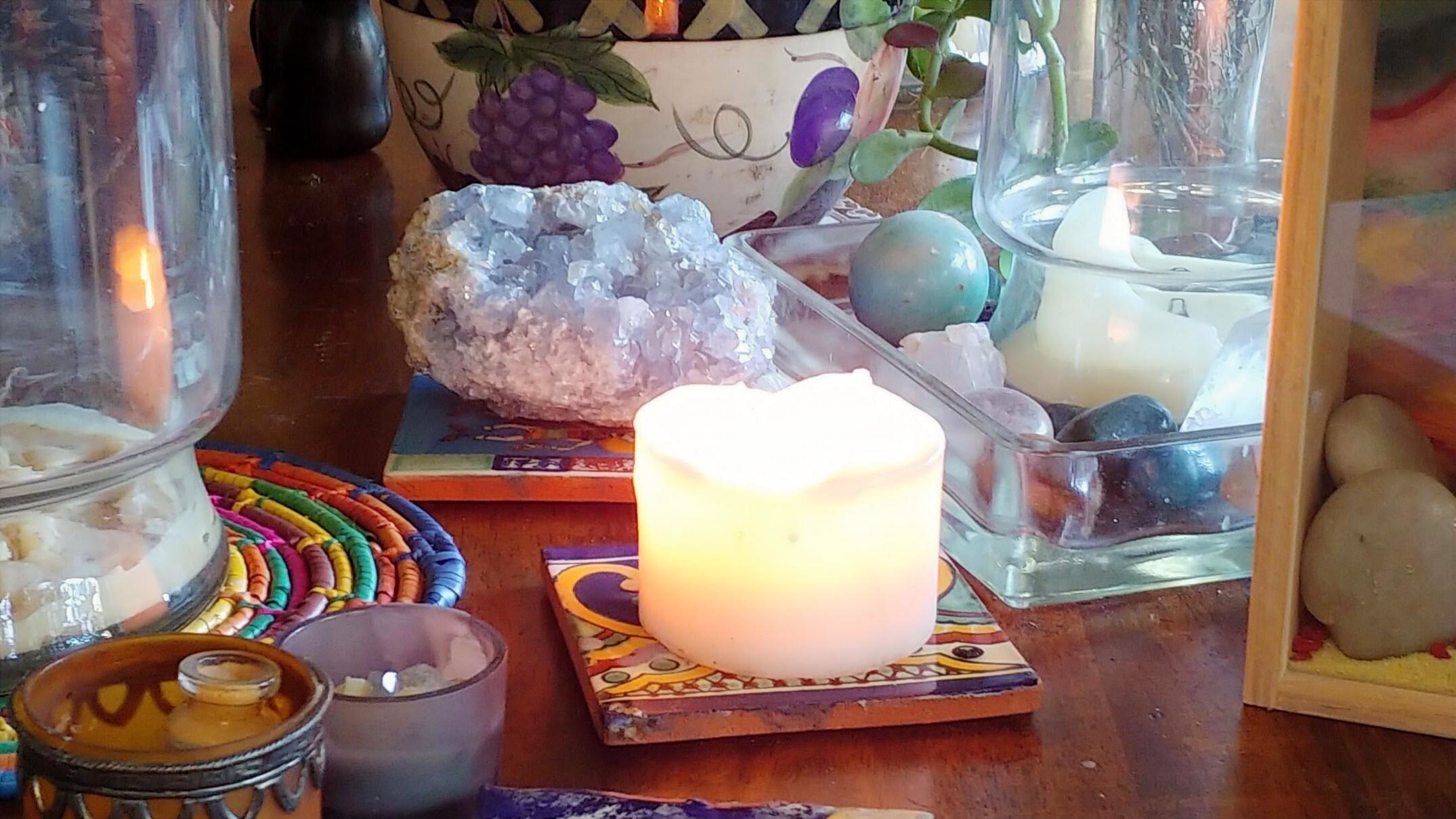 A lit candle on a hand-painted tile, surrounded by stones, a glass jar, and warm objects on a wooden surface