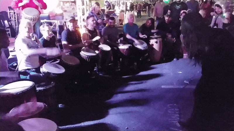 A drum circle in motion — many hands on hand drums, bodies leaning into the rhythm, a circle of people held together in shared beat