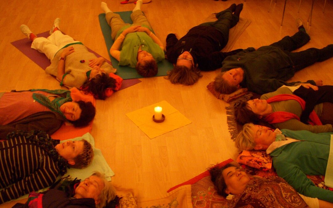 A group of people lying on the floor in a circle, feet pointed inward toward a central candle and in the integration phase of a breathwork session