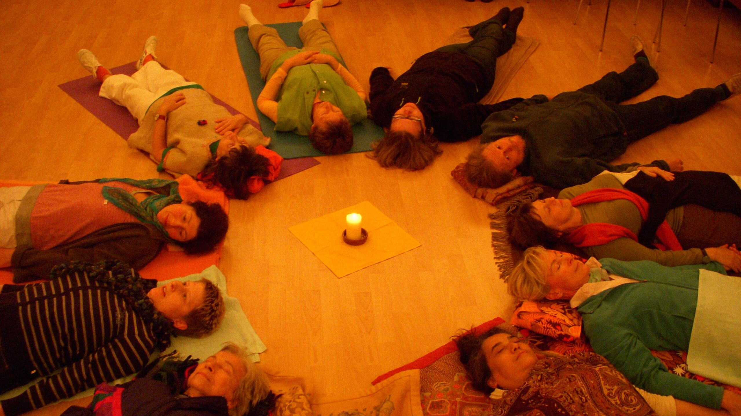 A group of people lying on the floor in a circle, feet pointed inward toward a central candle and in the integration phase of a breathwork session